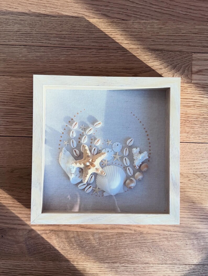 May include: A framed beach-themed artwork featuring a collection of seashells, starfish, and coral arranged on a light-colored fabric. The arrangement is enclosed in a square, light-wood frame, creating a coastal aesthetic. The artwork is displayed on a wooden surface.