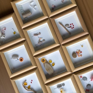 May include: A collection of nine square light wood frames containing pebble art. Each frame features small smooth stones arranged to form figures, birds, or flowers, accented with twigs, painted details, and small colorful floral embellishments.
