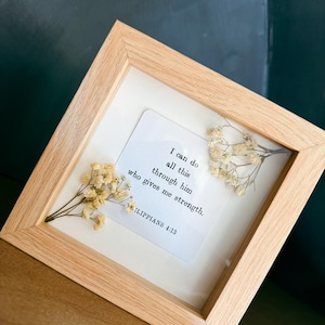 Könnte beinhalten: Ein quadratischer, heller Holzrahmen mit einer weißen Karte, auf der in schwarzer Schrift steht: I can do all this through him who gives me strength. PHILIPPIANS 4:13. Zwei Zweige getrockneter, cremefarbener Schleierkrautblüten sind darin gepresst.