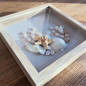 May include: A shadowbox frame with a light wood finish, showcasing a collection of seashells and a starfish on a neutral linen background. The arrangement includes various shells, small starfish, and coral, creating a coastal theme.