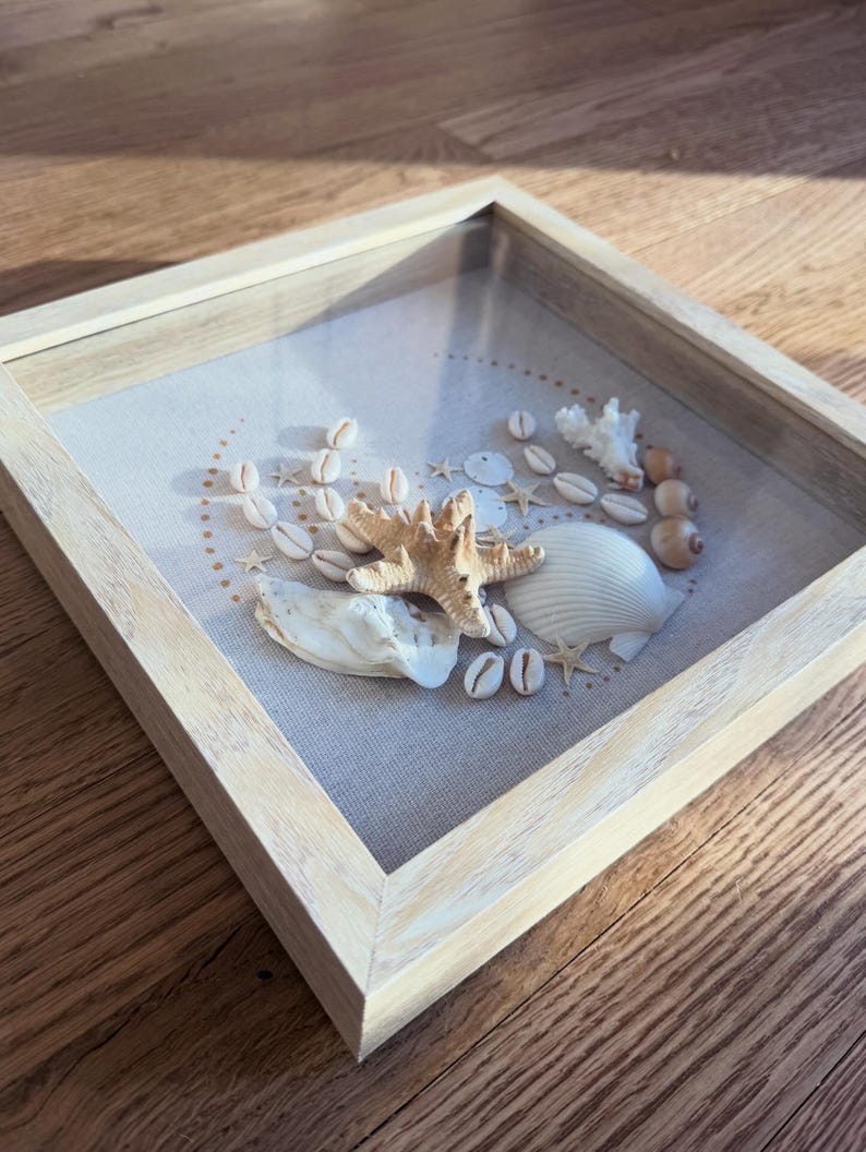 May include: A shadowbox frame with a light wood finish, showcasing a collection of seashells and a starfish on a neutral linen background. The arrangement includes various shells, small starfish, and coral, creating a coastal theme.