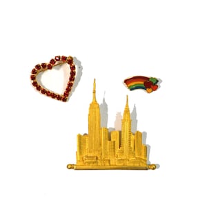 May include: A gold-colored pin featuring a stylized cityscape with a rainbow pin above it and a red rhinestone heart pin to the left.