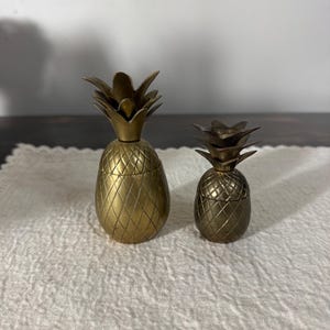 May include: Two brass pineapple-shaped ornaments. The larger one is approximately 13 cm tall, and the smaller one is about 8 cm tall. Both have a textured, diamond-patterned body and a leafy crown. They sit on a white, textured surface.