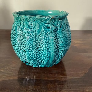 May include: A turquoise ceramic planter with a textured, dotted surface and a ruffled rim. The planter has a bulbous shape and is decorated with leaf and vine patterns. The color is a vibrant teal, suitable for indoor or outdoor use.