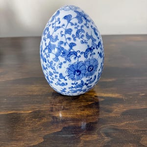 May include: A decorative ceramic egg with a blue and white floral pattern. The egg-shaped ornament features a detailed design of blue flowers and vines against a white background. The egg is displayed on a dark wooden surface.