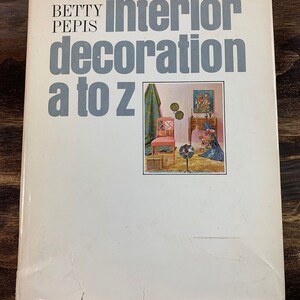 May include: A vintage book titled "Interior Decoration A to Z" by Betty Pepis. The cover is white with gray text and a small image of a room with furniture. The book shows signs of wear.