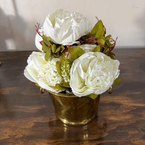 May include: A bouquet of white peonies with green leaves and red stems in a small, antique brass bucket. The flowers are clustered together, creating a full and lush arrangement. The bucket has two small handles.