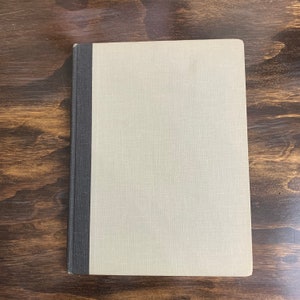 May include: A closed book with a light beige cover and a dark gray spine. The book is lying flat on a wooden surface. The cover has a textured appearance.