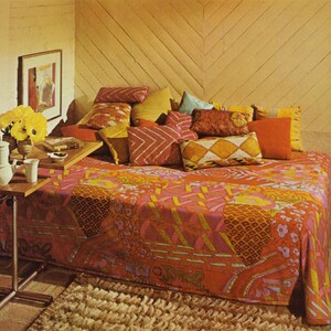 May include: A bed with a vibrant, patterned bedspread in shades of orange, pink, and yellow. Several decorative pillows in various colors and patterns are arranged against a wooden wall. A small table holds a coffee pot, cup, and flowers.