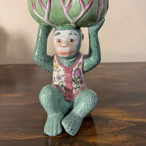 May include: A ceramic monkey figurine in a seated position, holding a green bowl above its head. The monkey is painted in shades of green, white, and pink, with a vest adorned with floral patterns. The bowl has a unique design with pink accents.