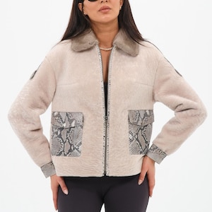 Könnte beinhalten: Beige Jacke mit Pelzkragen, Schlangenmuster-Taschen und Manschetten. Die Jacke hat einen Reißverschluss und wird mit schwarzen Leggings getragen. Das Model trägt eine Sonnenbrille.
