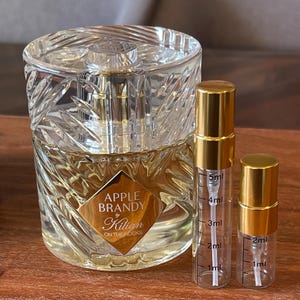 Puede incluir: Un frasco de perfume de vidrio transparente con un diseño tallado en diamante, lleno de un líquido ámbar claro. La botella está etiquetada como "APPLE BRANDY by Kilian ON THE ROCKS." Dos viales más pequeños con tapas doradas.