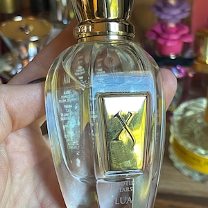 Könnte beinhalten: Eine transparente Parfümflasche mit goldenem Verschluss und einem rechteckigen goldenen Etikett. Das Etikett zeigt ein goldenes 'X'-Design. Der Text 'SHOOTING STARS LUA' ist auf der Flasche aufgedruckt. Die Flasche wird in einer Hand gehalten.