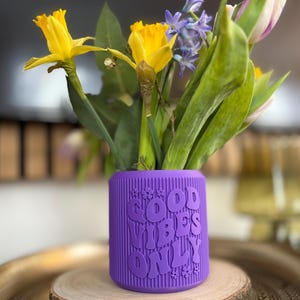 Könnte beinhalten: Eine lila Vase mit dem Schriftzug "GOOD VIBES ONLY" enthält einen Blumenstrauß. Gelbe Narzissen, violette Hyazinthen und weiße und rosa Tulpen sind zu sehen. Die Vase ist zylindrisch und geriffelt.