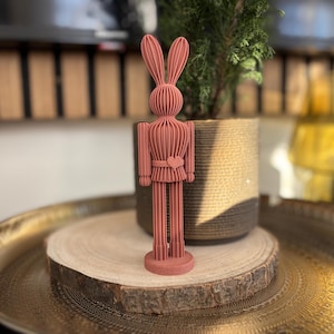 Puede incluir: Una figura de conejo de madera de color coral con orejas largas, una hebilla de cinturón en forma de corazón y una base redonda. El conejo está sobre una rodaja de madera, con una planta en una maceta tejida detrás. La figura está en una bandeja dorada.