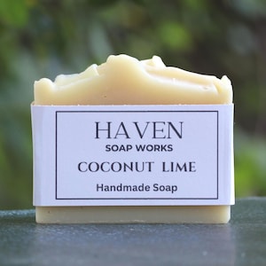 May include: A rectangular bar of off-white soap with a textured top. A white paper label is wrapped around the center, featuring the black text: HAVEN SOAP WORKS, COCONUT LIME, and Handmade Soap, enclosed within a thin black rectangular border.