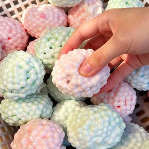 Puede incluir: Una mano sostiene un objeto esférico rosa claro con una superficie texturizada en forma de burbujas. El objeto está rodeado de esferas similares en tonos de rosa, verde y azul, todo dentro de una cesta blanca. Las esferas parecen ser jabón o bombas de baño.