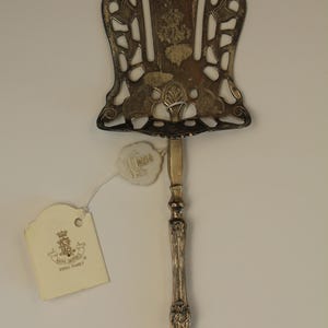 Può includere: Utensile da portata argentato con un design traforato ornato. Il manico è rifinito con motivi decorativi. Un'etichetta con le parole "Royal Butler" e un stemma è attaccata. L'utensile è probabilmente per servire torte o pasticcini.