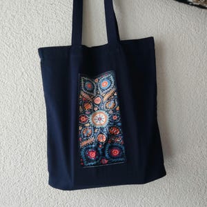 May include: Navy blue tote bag with a colorful, patterned patch. The patch features a detailed floral design with red, orange, blue, and yellow accents. The bag has two long straps for carrying over the shoulder.