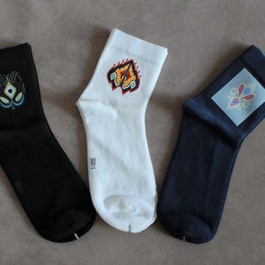 Könnte beinhalten: Drei Paar Socken mit einzigartigen Designs. Eine schwarze Socke mit einem teal-gelben Grafik, eine weiße Socke mit einem rot-orangen Design und eine marinefarbene Socke mit einem blau-roten Muster. Die Socken sind aus weichem Material.