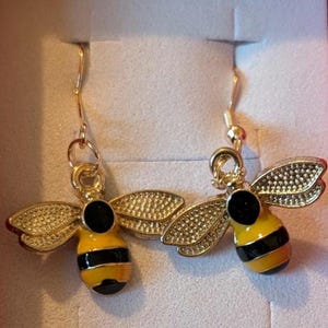 May include: A pair of gold-colored bee earrings. Each earring features a yellow and black striped body, black head, and textured gold wings. The earrings hang from gold-colored hooks.