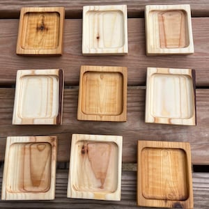 Könnte beinhalten: Neun quadratische Holztabletts mit vertieftem Zentrum. Die Tabletts bestehen aus hellem Holz mit sichtbaren Maserungsmustern. Einige Tabletts haben dunklere Kanten, und die Holztöne variieren von hell- bis mittelbraun. Die Tabletts sind auf einer Holzoberfläche angeordnet.