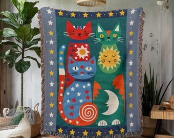 Puede incluir: Una manta turquesa con tres gatos estilizados con diseños de sol y luna. Los gatos son rojos, verdes y azules con patrones de estrellas. La manta tiene un borde azul con detalles de estrellas y bordes con flecos.