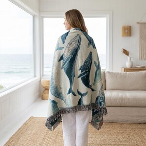 May include: A cream-colored throw blanket featuring a pattern of blue whales. The blanket has a fringed edge and is draped over a person. The background includes a window with an ocean view and a neutral-toned interior.
