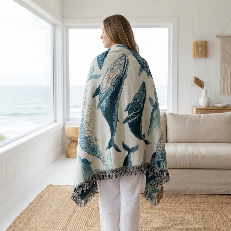 May include: A cream-colored throw blanket featuring a pattern of blue whales. The blanket has a fringed edge and is draped over a person. The background includes a window with an ocean view and a neutral-toned interior.