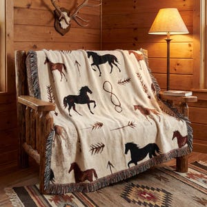 May include: A cream-coloured throw blanket featuring a horse and arrow pattern. The blanket is draped over a wooden bench. The horses are brown and black. The blanket has a fringed edge and is on a patterned rug.