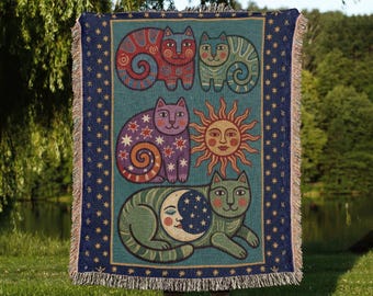Puede incluir: Una manta de color turquesa con ilustraciones caprichosas de gatos en rojo, verde y morado, junto con un diseño de sol y luna. La manta tiene un borde azul marino con patrones de estrellas y bordes con flecos.