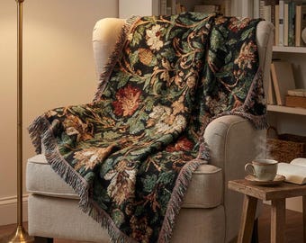 May include: A floral tapestry throw blanket draped over a beige armchair. The blanket features a dark background with red, green, and tan floral patterns. The edges are fringed. A floor lamp and small wooden side table are also visible.