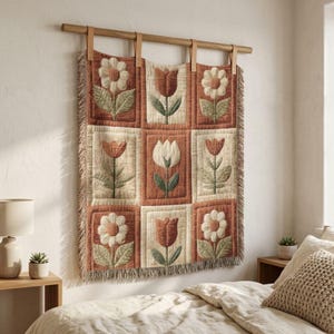 May include: A floral wall hanging with a grid pattern of nine squares. Each square features a different flower design in shades of cream, orange, and green. The hanging is mounted on a wooden rod with leather straps and has a fringed edge.