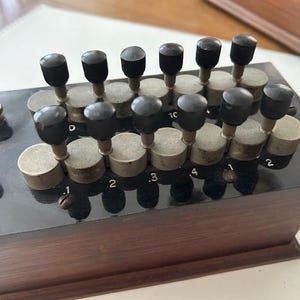Puede incluir: Un componente eléctrico vintage con una base de madera oscura y una parte superior negra brillante. Cuenta con múltiples perillas cilíndricas con tapas negras y diales numerados, probablemente utilizados para ajustar la resistencia eléctrica. El diseño sugiere un equipo clásico, posiblemente antiguo.