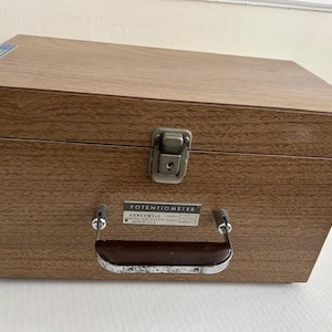 May include: A vintage wooden box with a metal latch and handle. The box has a brown finish and a label that reads "POTENTIOMETER" and "HONEYWELL". The box is likely used for storing or transporting electronic equipment.