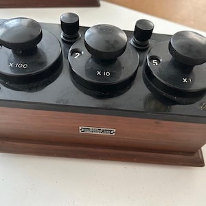 Puede incluir: Un componente eléctrico antiguo con base de madera y perillas negras. Las perillas están etiquetadas con números y multiplicadores, incluyendo "X 100", "X 10" y "X 1". El dispositivo tiene forma rectangular y una placa metálica con texto.