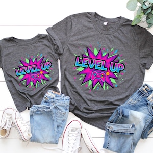 May include: Two gray t-shirts with a "LEVEL UP" graphic featuring a pixelated controller. The design is set against a burst of pink, green, and blue. The shirts are paired with blue jeans and white sneakers.