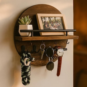 May include: A circular, dark wood wall shelf with a small shelf, hooks, and a black metal bar. It displays a framed photo, a small potted succulent, keys, sunglasses, and a watch. The shelf is mounted on a white wall.