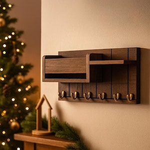 Pode incluir: Um organizador de parede de madeira castanha escura com uma prateleira, uma ranhura para correspondência e seis ganchos de cor latão. O organizador é montado numa parede bege clara. O design é simples e moderno, adequado para a entrada ou organização doméstica.