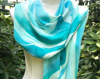 Puede incluir: Bufanda fluida con diseño de acuarela en tonos turquesa, verde azulado y blanco. La bufanda está drapeada sobre un maniquí, mostrando su textura suave y su vibrante paleta de colores. El diseño de la bufanda presenta pinceladas abstractas.
