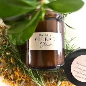 May include: A brown glass jar labeled "BALM of GILEAD Glow" with a black lid, surrounded by dried herbs and greenery. The lid has a white label with a quote and a wheat illustration. The image is a close-up shot.