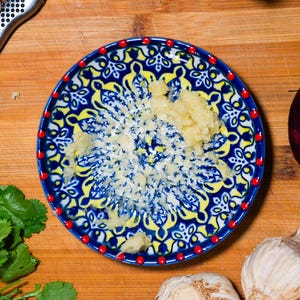 Peut inclure: Un presse-ail en céramique bleu et jaune avec un bord à points rouges, rempli d'ail râpé. Un couteau en acier inoxydable, un oignon rouge, de la coriandre et une gousse d'ail sont également visibles sur une planche à découper en bois. Outils culinaires.