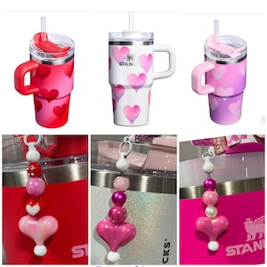 Könnte beinhalten: Drei isolierte Edelstahlbecher mit Herzmustern in Rot, Weiß und Rosa. Jeder Becher hat einen Griff, einen Deckel mit Strohhalm und einen Perlenanhänger. Die Anhänger zeigen rosa und weiße Perlen und einen herzförmigen Anhänger.
