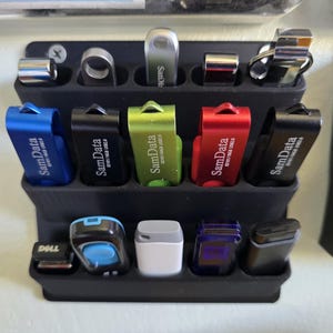 May include: A black plastic tiered storage rack holding fourteen USB flash drives of various colors and designs. The drives include blue, black, green, red, silver, and white models, with some labeled SamData and one labeled Dell, arranged in three rows.