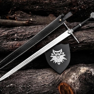 Puede incluir: Una espada plateada con empuñadura y guarda de color negro, sobre una superficie de madera. También hay una vaina de cuero negro y un escudo negro con un emblema de lobo. La hoja de la espada mide unos 91 cm.
