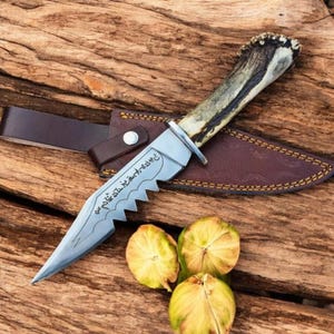 May include: A fixed-blade knife with a textured antler handle and a silver-toned steel blade featuring a serrated spine and etched runic-style inscriptions. The knife is accompanied by a brown leather sheath with visible yellow stitching.
