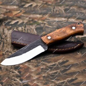 May include: A fixed-blade knife with a polished steel blade and a brown wooden handle secured by two silver rivets. The knife is positioned diagonally across a dark brown leather sheath featuring visible stitching along the curved edges.