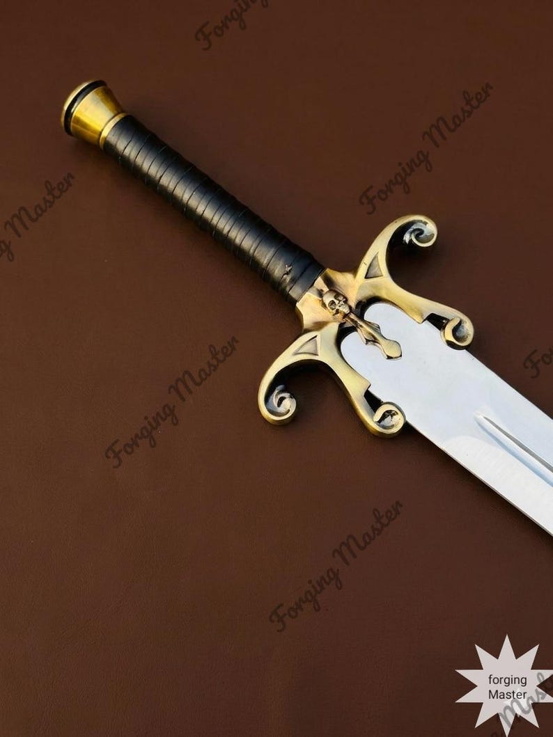 May include: A sword with a polished silver blade and a black wrapped handle. The hilt features a brass-colored crossguard with ornate, curved ends and a small skull emblem at the center. A brass-colored pommel caps the end of the handle.