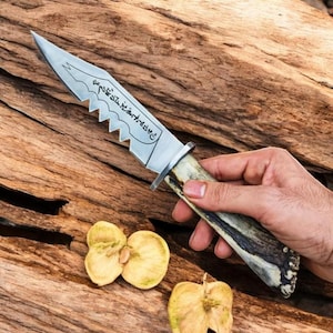 Può includere: Un coltello a lama fissa con una lama in metallo argentato seghettata che presenta scritte in nero incise. Il manico è realizzato in materiale testurizzato con tonalità marroni e crema, e il coltello ha una guardia in metallo argentato.