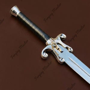 May include: A silver-toned sword featuring a black wrapped handle, a metallic pommel, and a crossguard detailed with a small skull emblem. The blade is polished steel with a central fuller, extending from a guard with curved, scroll-like quillons.